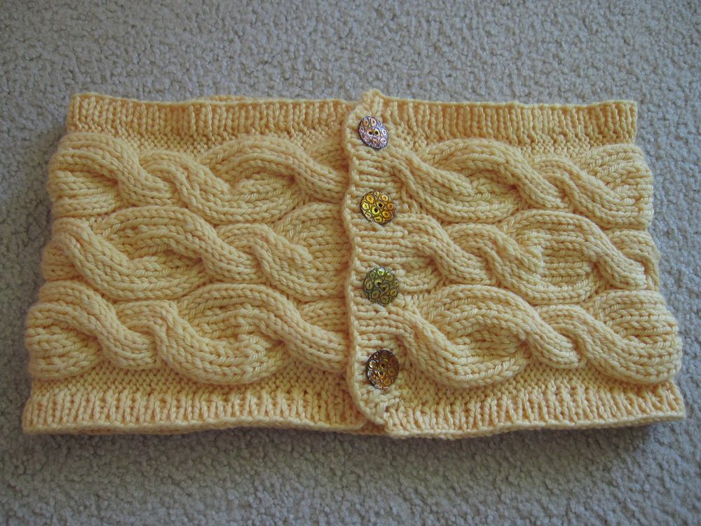 A photo of a pale yellow cowl lying flat on a neutral background. Hand-knit in a medium aran weight yarn, it is a wide but short loop that wraps around the neck once and fastens with a column of four round, flat, carved, gold-tinted mother-of-pearl buttons. The main body has three horizontal rows of closely-nestled cables that alternate thick and thin twists. The top and bottom edges, as well as the button band, have a thin border of ribbing.