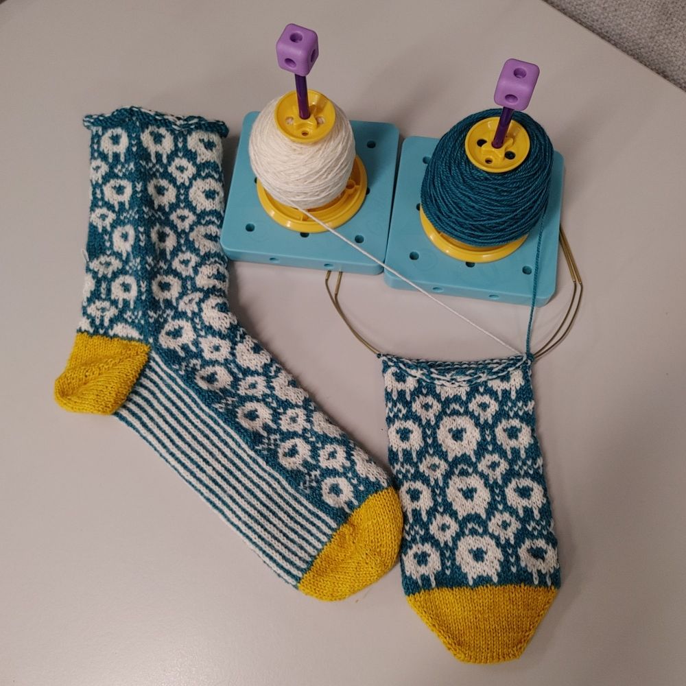An overhead view of a pair of handknit toe-up colorwork socks. One is almost finished except for the cuff. The other is still on the needles with the foot in progress. The main sock body is dark teal. They are dotted with white sheep and have pinstripe soles and yellow contrast toes and heels.