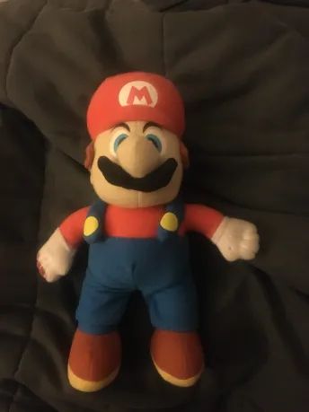 a photo of a kellytoy mario plushie laying on a gray blanket. he is looking at the viewer. there is red paint stains on his left hand and shoe.