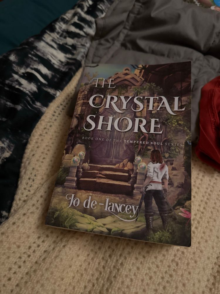 Jo’s novel in paperback form, lying on a bed. The cover has a mysterious dilapidated building with a person stood outside, back to the viewer. The person is masc presenting with a white shirt, long legs and wearing a substantial sword. 