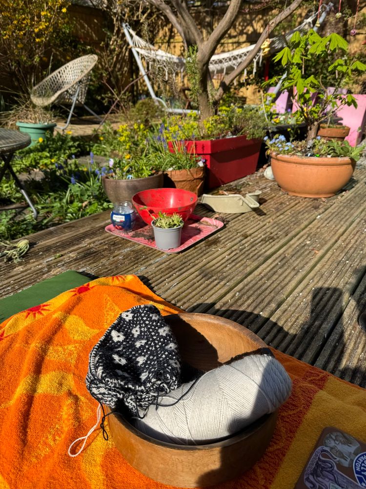 A photo of a messy Spring garden in morning sun. There is a hammock in the background. In the foreground is a wooden bowl with knitting- dark blue and white- a textured geometric knit. 