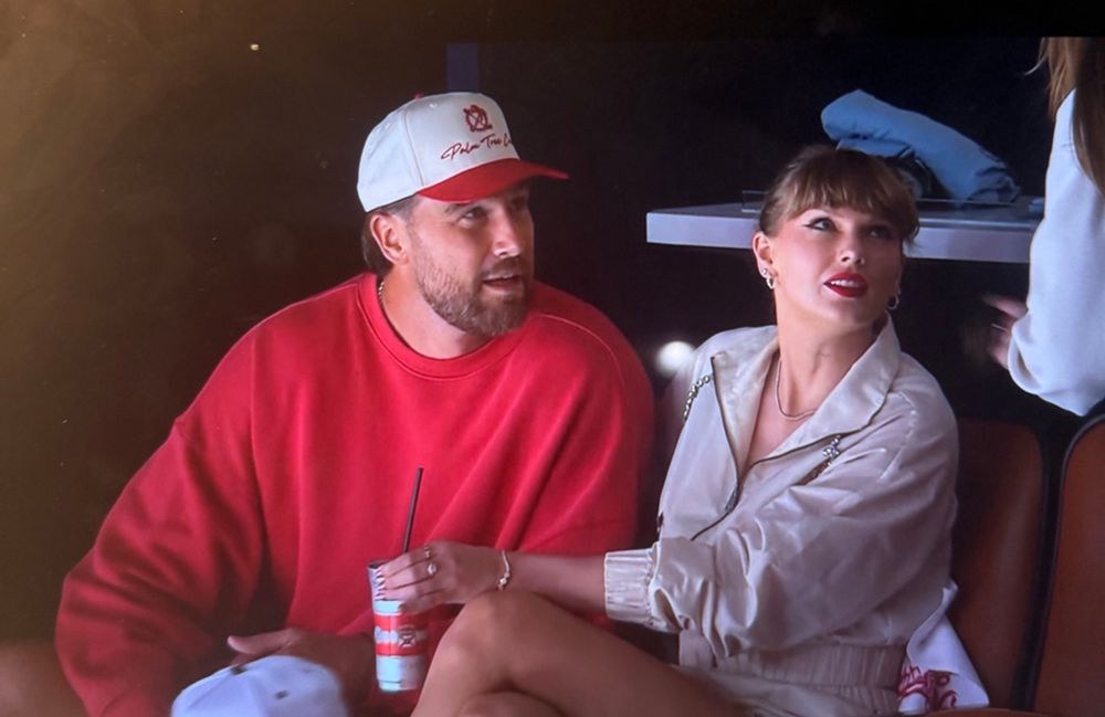 Travis kelce is on the left wearing a red sweater and a white hat. Taylor swift is sitting next to him gazing up. Her hair is back and she has red lipstick on