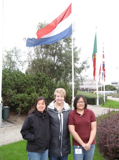 The same woman, about 12 years later. She stands outside, posing for an informal picture with two teenage girls. Behind them are some bushes, and a semicircle of different international flags. The photo was taken at the Baikonur Cosmodrome in 2007.