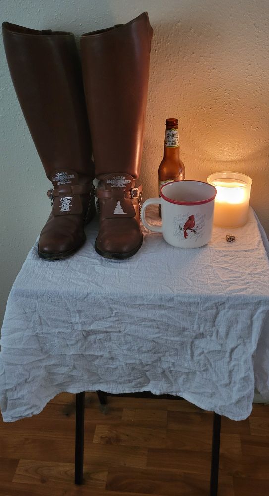 A small folding table covered in a white cloth, which holds a pair of knee high Corps of Cadets boots, a Shiner beer bottle, a mug, a lit white candle, and an Aggie ring arranged in tribute. 