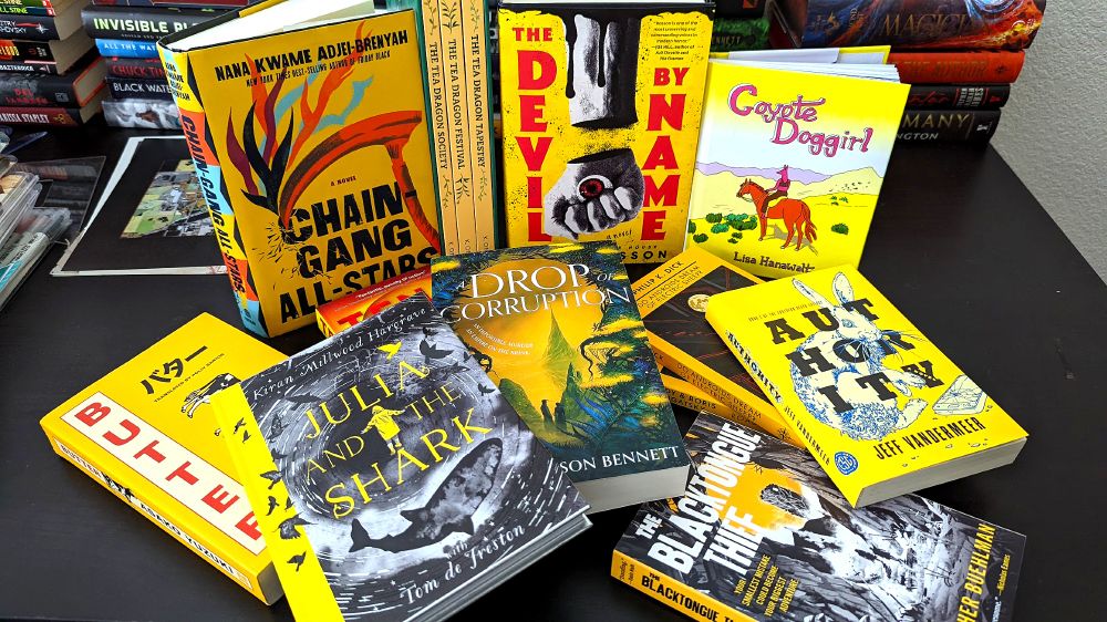 A pile of yellow books on a black tabletop with piles of other different colored books in the background. Yellow books featured include Butter by Asako Yuzuki, Chain-Gang All-Stars by Nana Kwame Adjei-Brenyah, Julia and the Shark by Kiran Millwood Hargrave, Ten Low by Stark Holborn, A Drop of Corruption by Robert Jackson Bennett, The Devil By Name by Keith Rosson, Do Androids Dream of Electric Sheep? by Philip K. Dick, Roadside Picnic by Arkady and Boris Strugatsky, The Blacktongue Thief by Christopher Buehlman, Authority by Jeff VanderMeer, Coyote Doggirl by Lisa Hanawalt, and The Tea Dragon Society, The Tea Dragon Festival, and The Tea Dragon Tapestry by K. O'Neill.