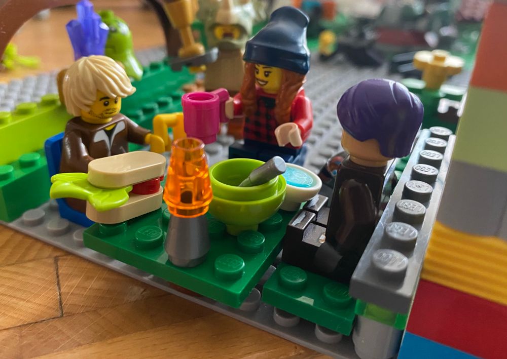 3 Lego-Püppchen sitzen an einem kleinen Tisch. Eine hat einen mittellange blonde Haare mit Seitenscheitel und trägt einen Stoppelbart und einen braunen Jogginganzug. Vor ihr stehen ein Sandwich mit Tomate und Salat sowie eine Lampe. In der hand hält sie einen gelben Becher. Zweite Figur trägt eine blaue Mütze über den langen dunklen Haaren und ein rotkariertes Hemd. In der Hand ein pinker Becher. Die letzte Figur ist dunkel gekleidet, hat lila Haare und vor ihr steht eine Schale mit Löffel.  Im Hintergrund sieht man verschwommen eine figur mit triceratops-maske und Schnauzbart, die eine Art Eisbecher trägt. 