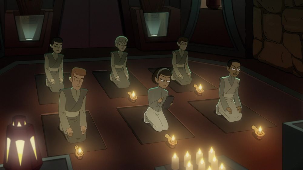 T’Lyn (center, forward) kneels on a meditation mat with a lamp in front of her doing work on a padd while most of the room is engaged in meditation