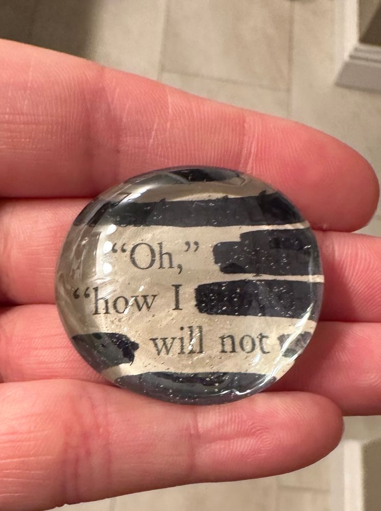 A largeish flat glass marble with a scrap of book page decoupaged to the back. Certain words have been blacked out and the remaining text says “Oh,” “how I will not