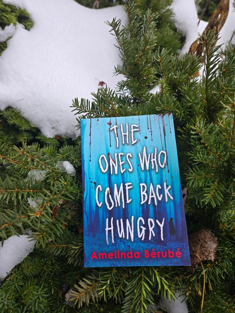 A copy of The Ones Who Come Back Hungry by Amelinda Berube on a background of evergreen branches and snow