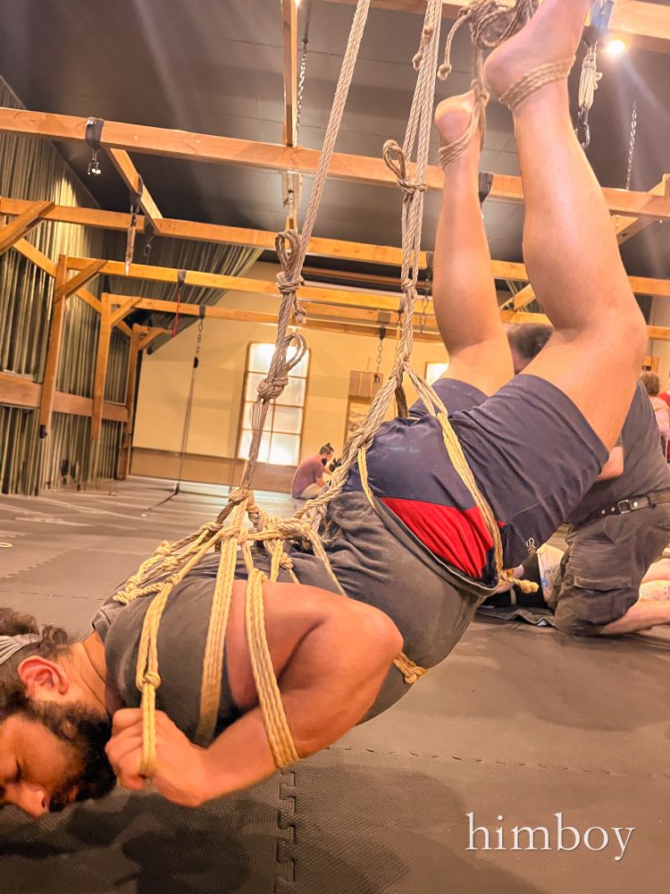 A brown non binary person with a beard suspended in shibari 