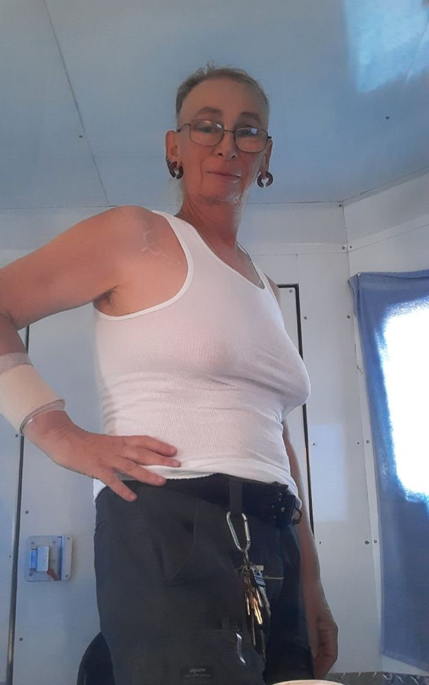 A trans woman with her hair tied back and sides shaved. She is wearing a men's white ribbed undershirt, and cargo shorts. She is sporting large purple glass hoop earrings, and wearing her keys on a carabinier.