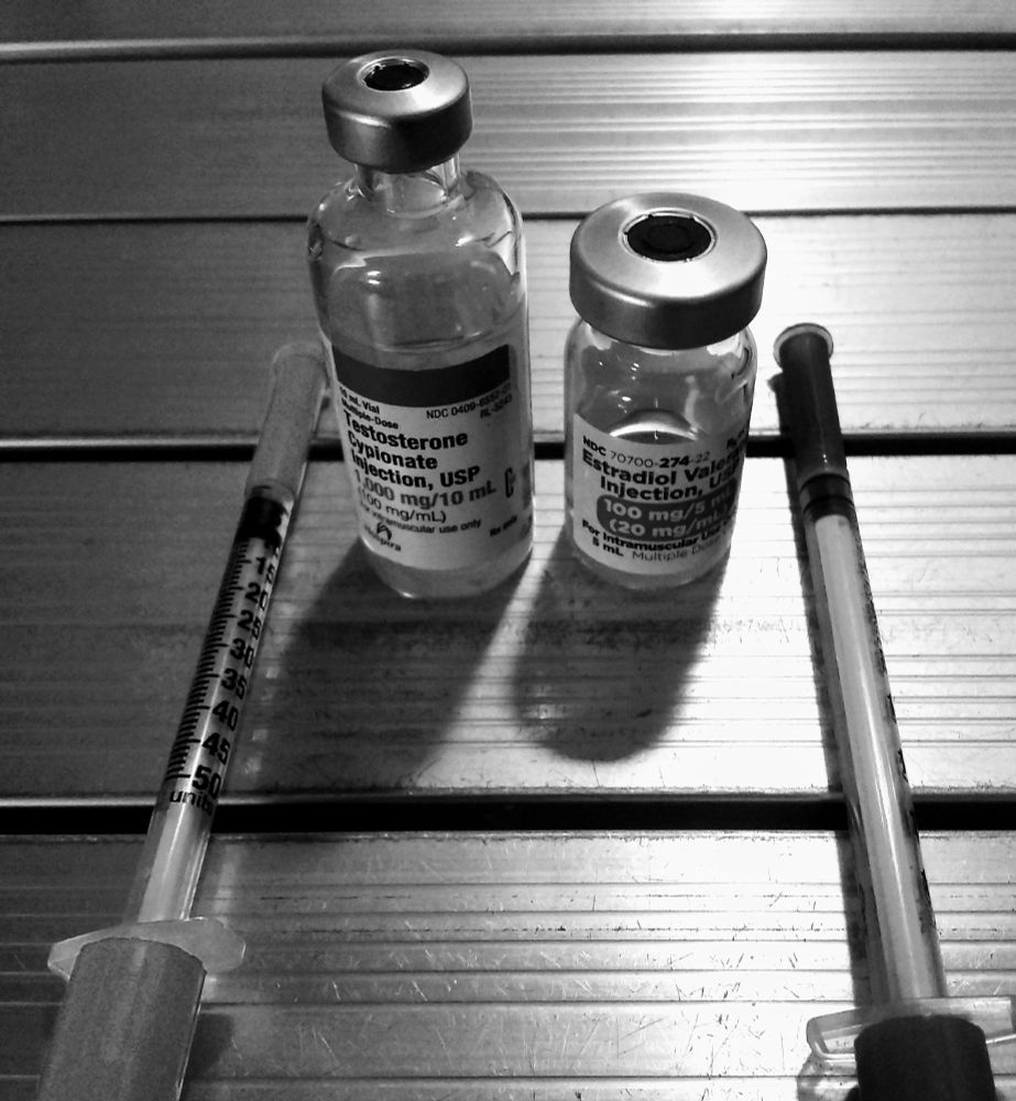 A table with two syringes, a vial of estradiol, and a vial of testosterone