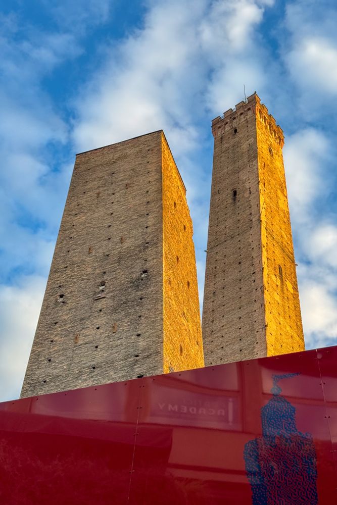 Das Bild zeigt zwei sehr hohe, alte Türme aus hellem Backstein, die in den Himmel ragen. Der linke Turm ist etwas breiter und wirkt schief, er neigt sich leicht zur Seite. Der rechte Turm ist schlanker, deutlich höher und hat oben eine gezackte Krone. Beide Türme sind in warmes Sonnenlicht getaucht, während darüber blauer Himmel mit weißen Wolken liegt.

Im Vordergrund sieht man ein rotes Baugerüst oder eine Absperrung, die aber nur einen kleinen Teil verdeckt. Die Türme selbst dominieren das Bild – sie wirken sehr massiv und eindrucksvoll.