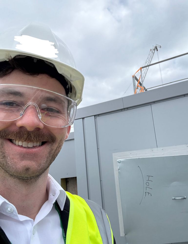 Selfie of Patrick wearing a hard hat - there’s a vent behind with the word HOLE written in 
marker 