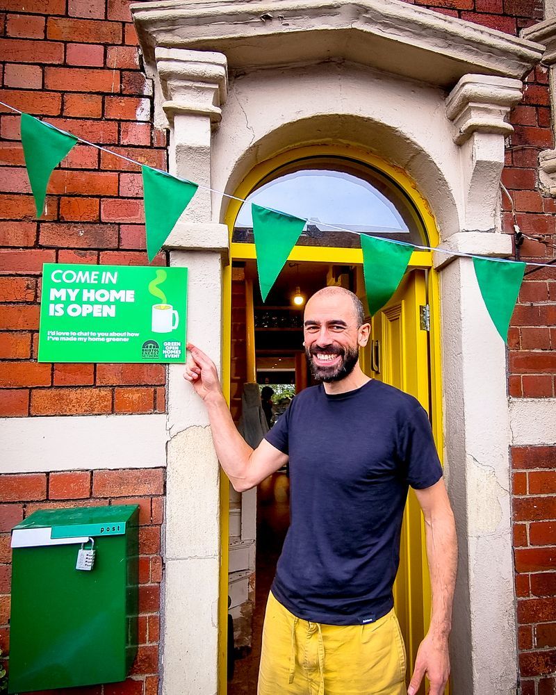 A smiling man pointing at a sign next to his door, it reads: Come in, my home is open.