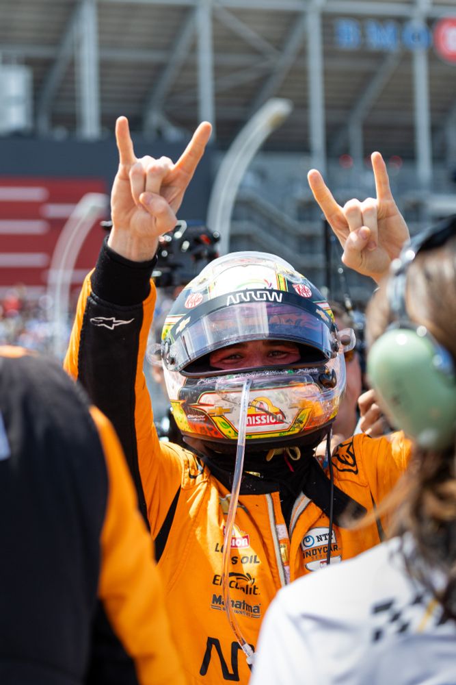 Pato O'Ward celebrating victory in Toronto on Sunday. He is wearing a helmet and orange racesuit. He is also doing the 🤟🏻 gesture. 