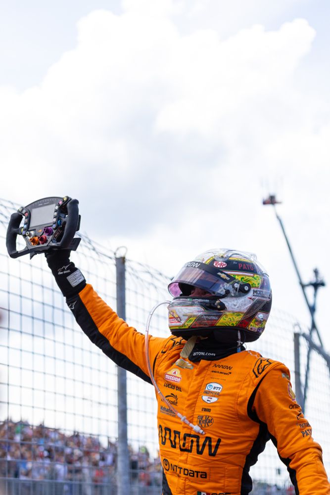 Pato O'Ward celebrating victory in Toronto on Sunday. He is wearing a helmet and orange racesuit. He is also holding a steering wheel in his right hand as he salutes the crowd. 