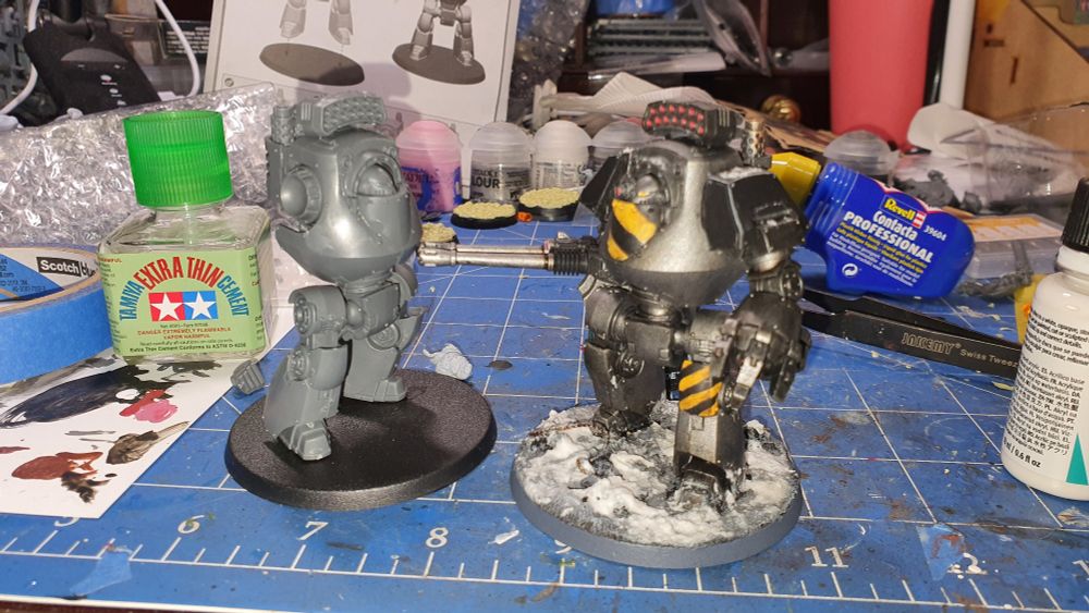 2 contemptor dreadnoughts on a messy work bench. One unpainted being held up while the glue drys by the painted one.