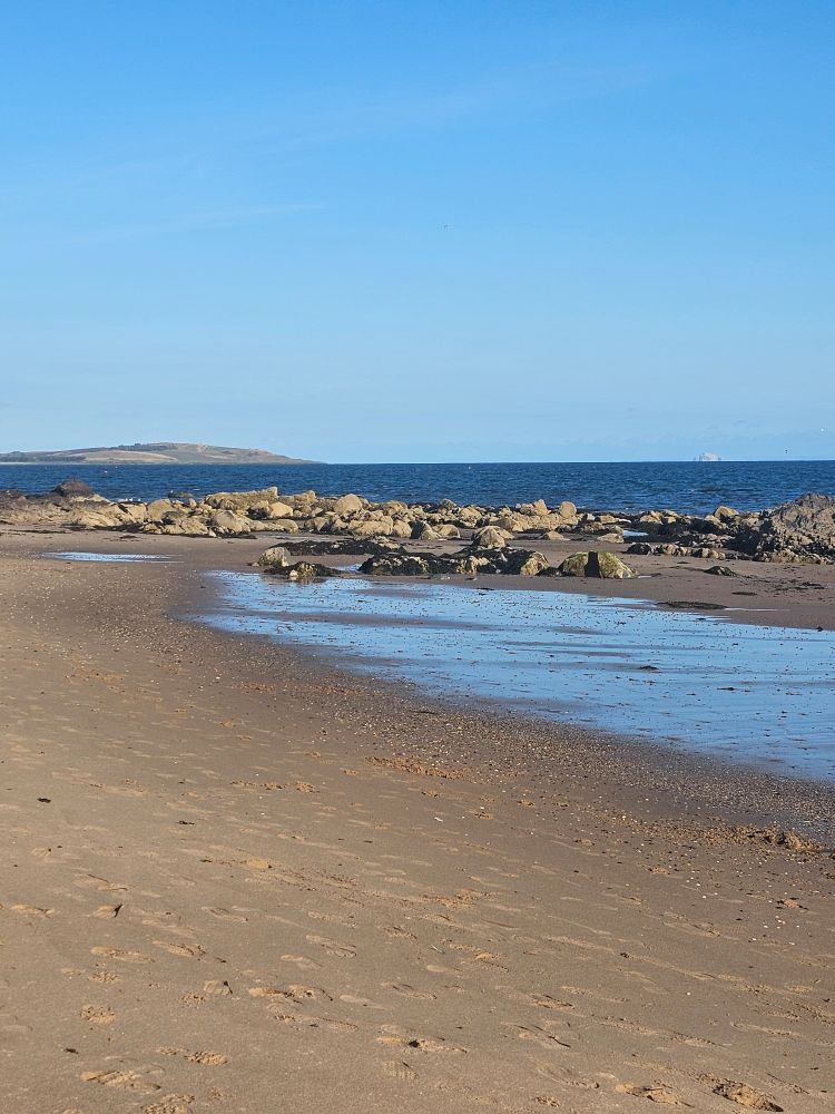 Ludin Links beach. The sky is very blue and the sea is darker but still bright. The wet sand looks like a mirror with the sunlit rocks providing a gilt frame. 