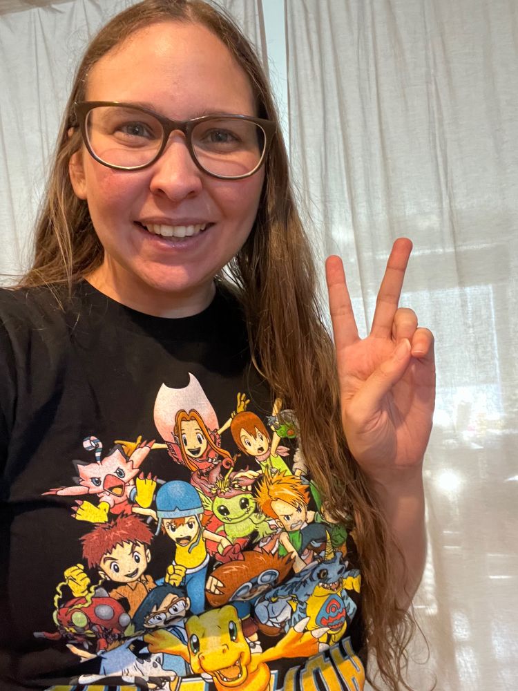 A brown haired woman with glasses is smiling at the camera while my asking a peace sign. She’s wearing a black T-shirt with various characters from the anime digimon on it.