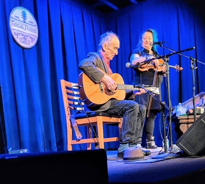 English folk musicians Martin Carthy and his daughter Eliza Carthy on stage at the Focal Point in Maplewood, Missouri. Martin is sitting on a wooden chair with an acoustic guitar on his knee and his hat is on the floor by his feet. Eliza stands to his left with her violin, smiling at Martin while speaking into a microphone. Behind them is a blue curtain and the Focal Point sign. 
