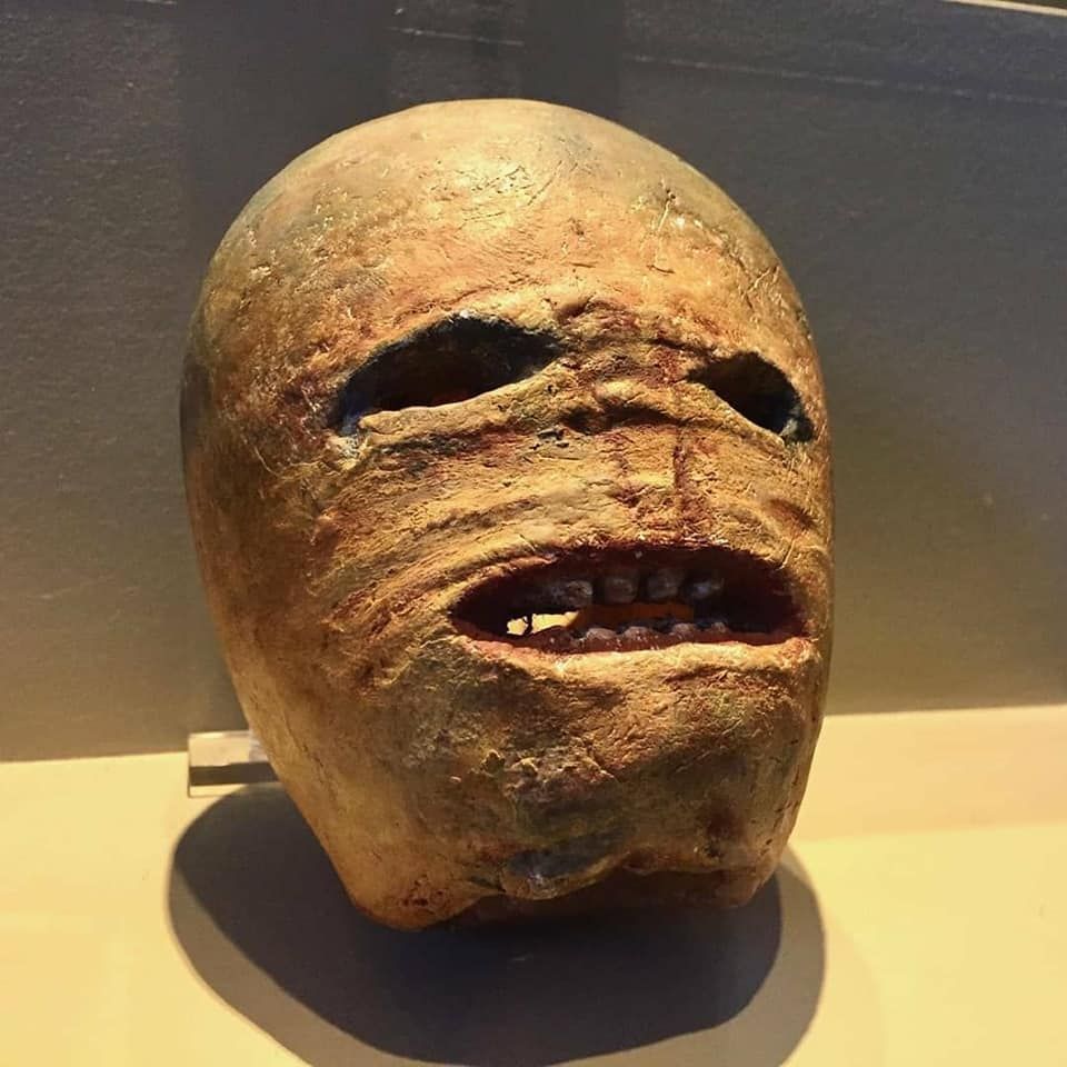 A traditional carved turnip lantern for Samhain, this one is in the Museum of Country Life in county Mayo. This one has wide, empty eye sockets and a wide, open mouth showing a few small, irregular teeth. The face appears wrinkled and distorted in a grimace or look of terror. It's on a brown base.