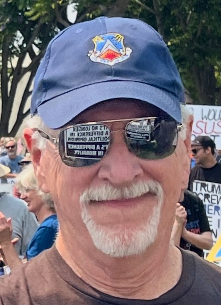 White old guy with grey goatee and aviator sunglasses wearing a blue ball cap smirking at camera