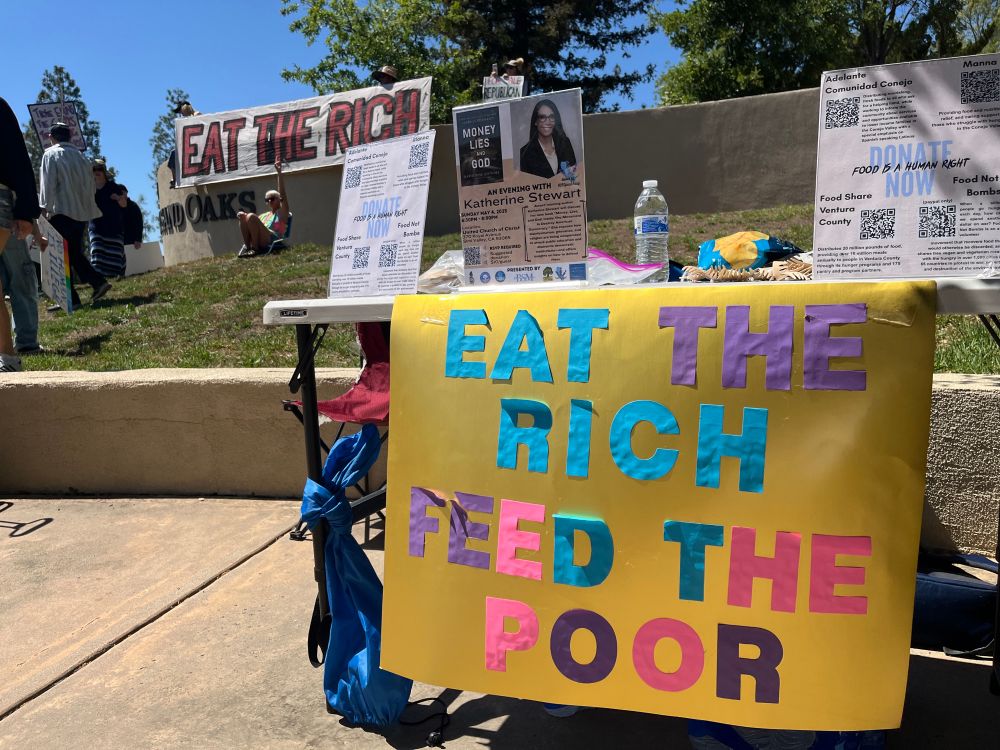 Photo of a table with a sign attached that reads “eat the rich feed the poor”. There are flyers on the table with QR codes linked to donations for local orgs. In the background demonstrators display a large “eat the rich” banner.