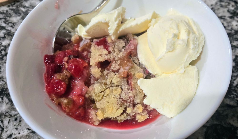 A bowl of cherry rhubarb crisp and scoops of vanilla ice cream with a spoon in the upper left.