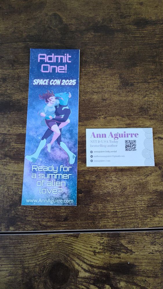 Bookmark: 

starry galaxy in background in purples, blues and pinks
Admit one! in purple font
Space Con 2025 (white font)

(cartoon style illustration) Blue dragonish alien is hug-carrying our human heroine, a chubby woman with a brown ponytail. She looks so happy.
Bottom of bookmark:

Ready for a summer of alien love? (white font)
www.annaguirre.com (white font)

The business card is white with lavender circles on the right side, it says Ann Aguirre NYT & USA Today bestselling author (darker purple)

annaguirre.bsky.social
 authorannaguirre@gmail.com
annaguirre.com

There's also a QR code to join my discord.