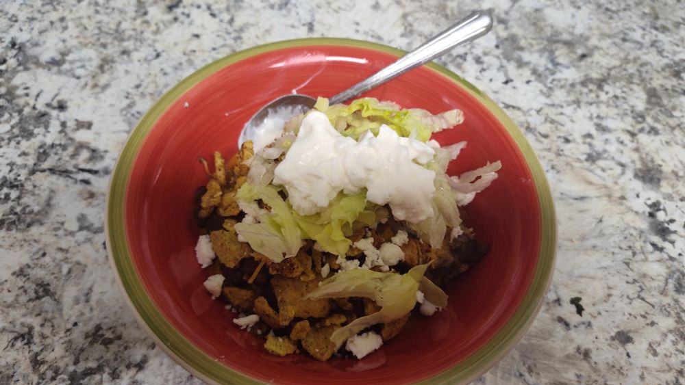 A red and green fiesta ceramic bowl full of chili enchilada casserole, topped with shredded lettuce, crumbled Panela cheese, and sour cream
