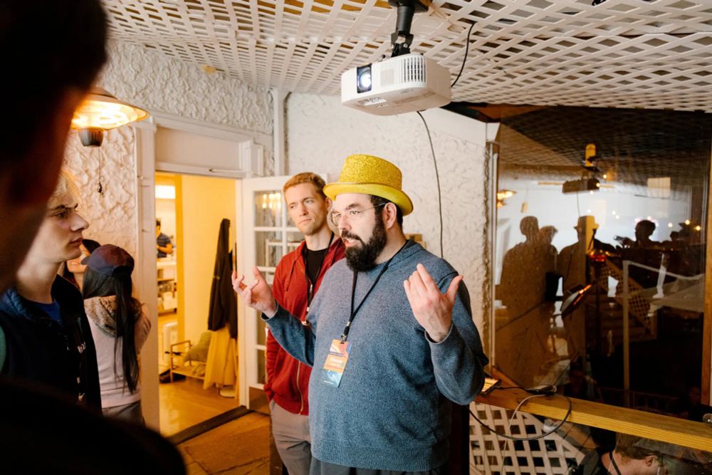 Rich white people staring at a husky white man with a dark beard wearing a gray sweater and a yellow hat