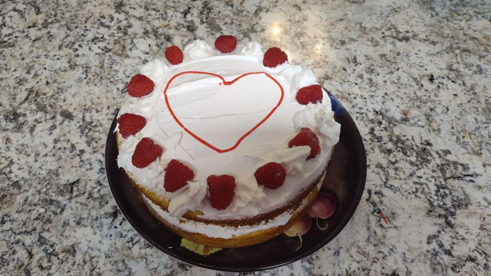 Raspberry lemonade cake It's a round layer cake covered in whipped cream layered with raspberries. The top has a red heart drawn on it and decorated with raspberries 