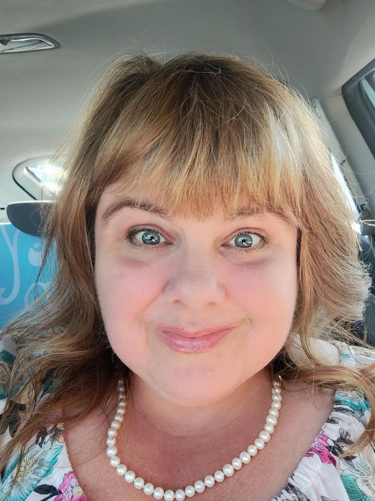 mildly sunkissed white lady with light brown / dark blond hair and gray kiss, wearing a white flowy dress with floral print and white pearls