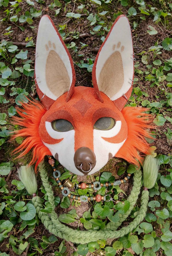 An image of an orange fox mask with large ears and green braids. It has green eyes, earrings, and a darker orange marking on its forehead in the shape of a 4 point star.
