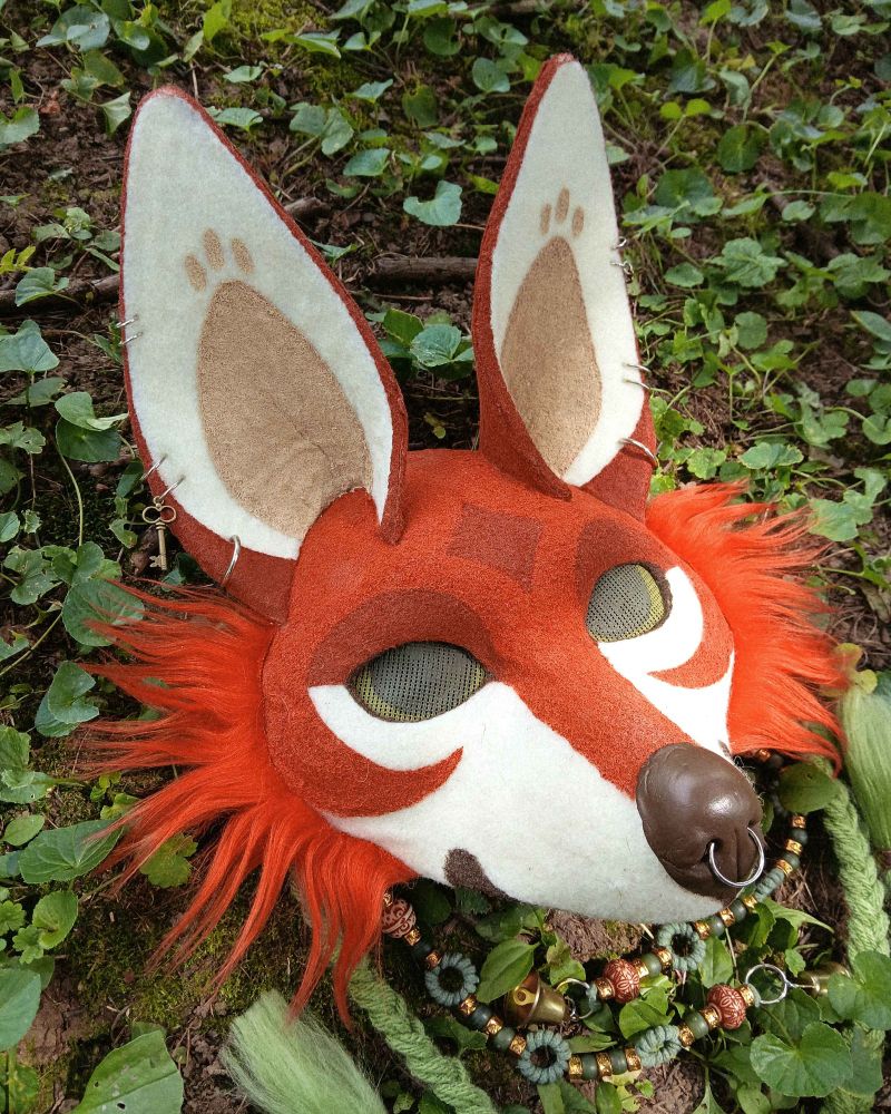 An image of an orange fox mask with large ears and green braids. It has green eyes, earrings, and a darker orange marking on its forehead in the shape of a 4 point star.
