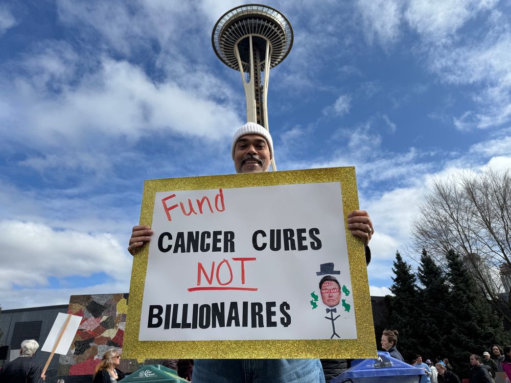 Me holding a sign that says “Fund cancer cures not billionaires”