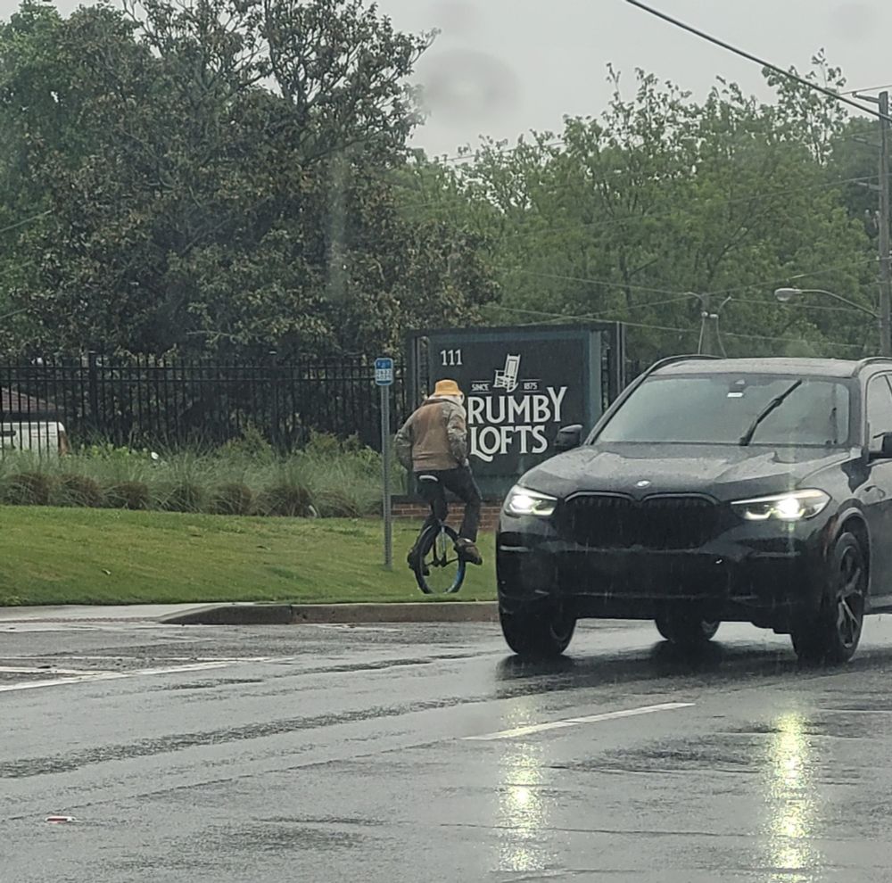 A man on a unicycle wearing a yellow hat riding it in the rain while a car passes in the foreground