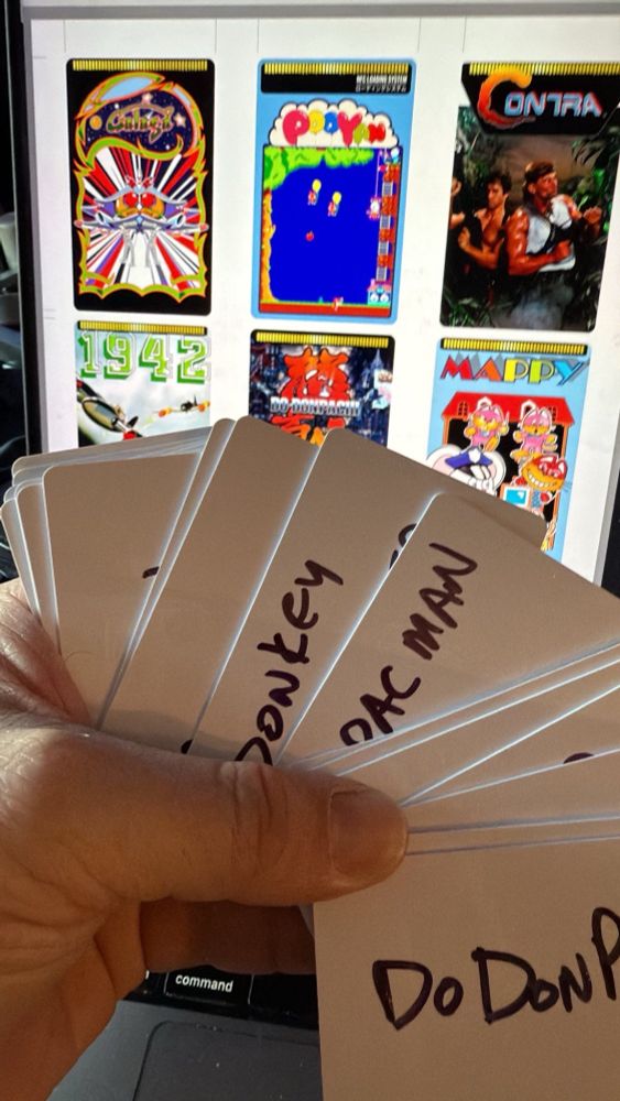 A hand holding a stack of white NFC cards, fanned like a deck of playing cards, with the names of video games on them: Donkey Kong, Ms Pac-Man and Do Donpachi are visible. Behind the cards, some graphic designs on a computer monitor for labels: Galaga, Pooyan, Contra, 1942, Do Donpachi and Mappy are visible.