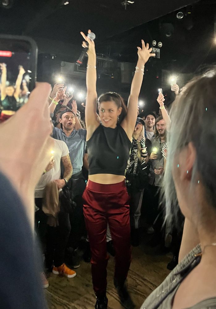 Rapper, writer, and vocalist Dessa performing at LPN in NYC in October 2023. At the end of the second encore, she is surrounded by fans illuminating the floor with their cell phone lights. Dessa stands with her arms raised and a huge smile on her face.