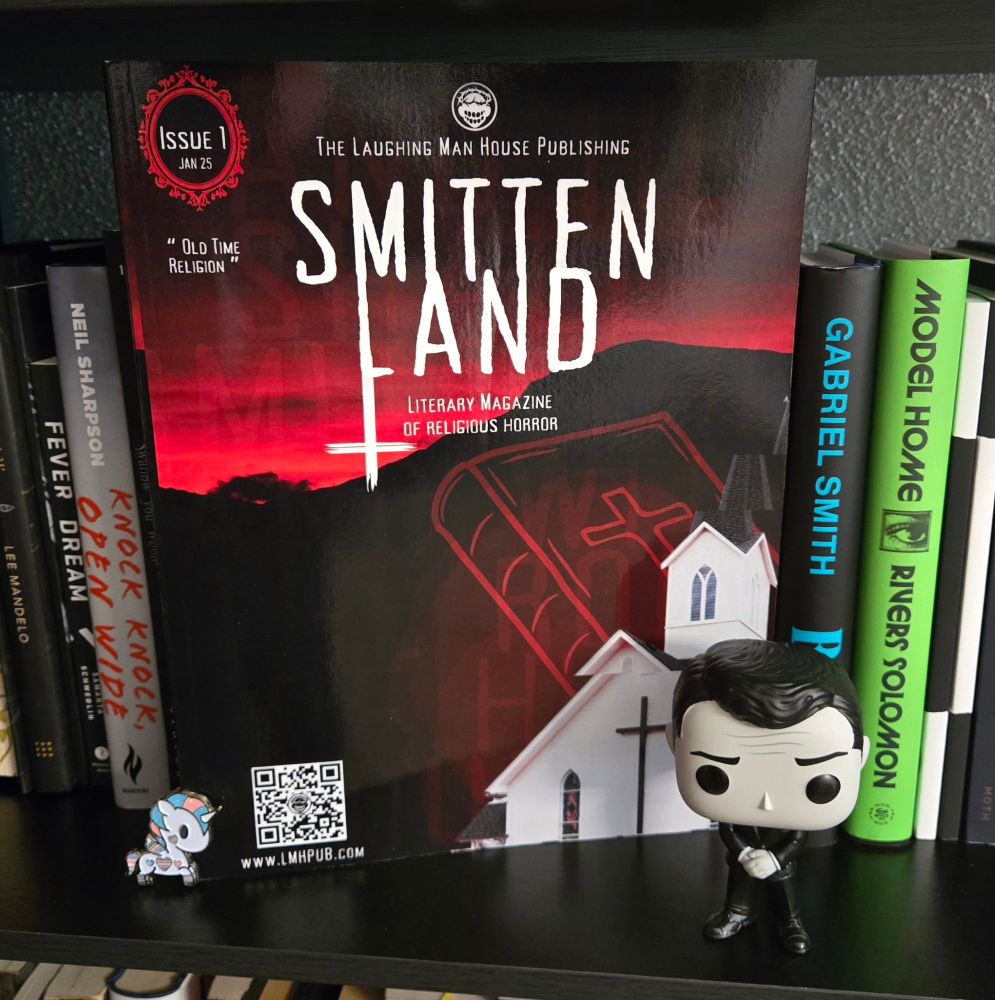 Copy of Smitten Land Literary Magazine of Religious Horror on a bookshelf. Flanked by a enamel pin of a Tokidoki unicorno with the trans flag colours and a Funko Pop of Rod Serling. 