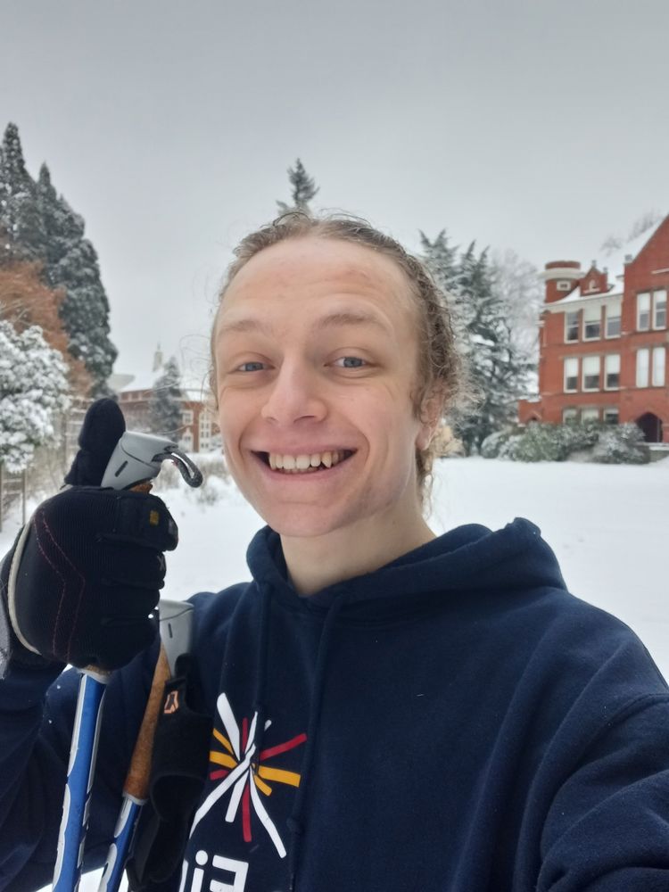 A photo of me, smilimg, holding a ski pole and giving a thumbs up to the camera. I'm white, with my brownish hair tied back. I'm wearing a navy blue hoodie and ski gloves. In the background are brick buildings on the Willamette University campus.