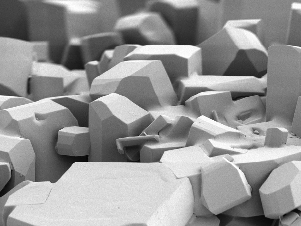 High-magnification scanning electron microscope (SEM) image of crystalline structures forming sharp, angular facets. The surfaces appear smooth and blocky, resembling a miniature abstract cityscape or tightly packed geometric shapes.