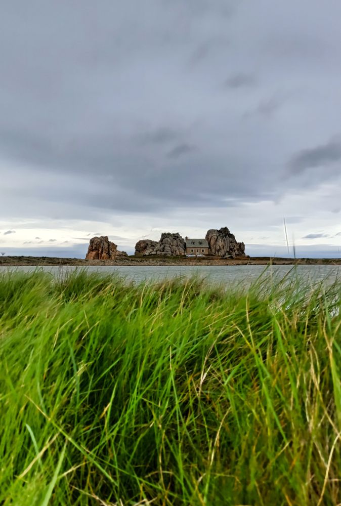 Maison encastrée entre deux gros rochers, avec la mer dans le dos et un étang devant. Deux voitures sont stationnées devant le bâtiment. Au premier plan, herbe balayée par le vent. Le ciel est nuageux et plus lumineux à l'horizon.