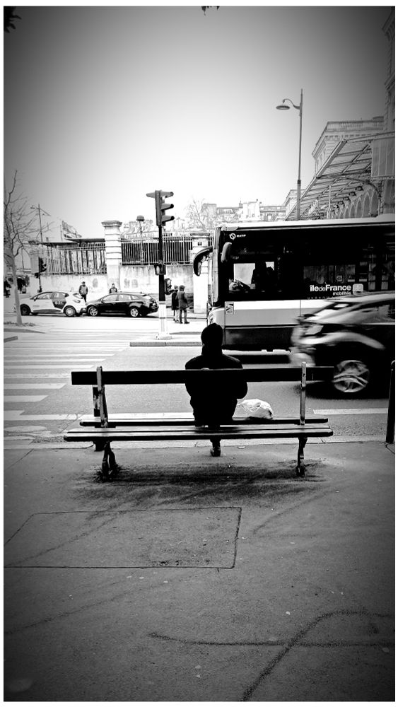 Photo en noir et blanc d'un homme assis sur un banc, habillé de sombre avec un bonnet sur la tête. Un sac en plastique blanc est posé à sa gauche. Dans la rue  où on voir un passage piéton ( à gauche) et un feu tricolore, un bus est à l'arrêt alors qu'une voiture disparaît sur la droite. En arrière plan, des passants été des voitures garées... Une grille entouré l'esplanade de la Gare de l'Est dont ont voit la structure métallique sur la droite. 