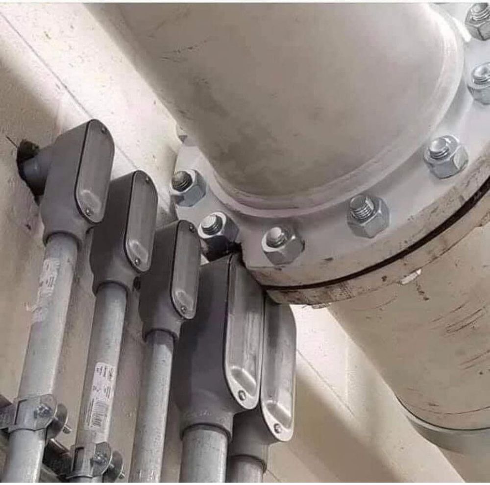 A large-diameter plumbing pipe with a wide, bolted-flange join runs along the ceiling. 

From the wall just below, several electrical conduits emerge.

A distressingly large chunk of the flange connecting the pipe sections has been ground away to make way for two of them, presumably compromising the strength of the seal and inviting all kinds of disaster. 
