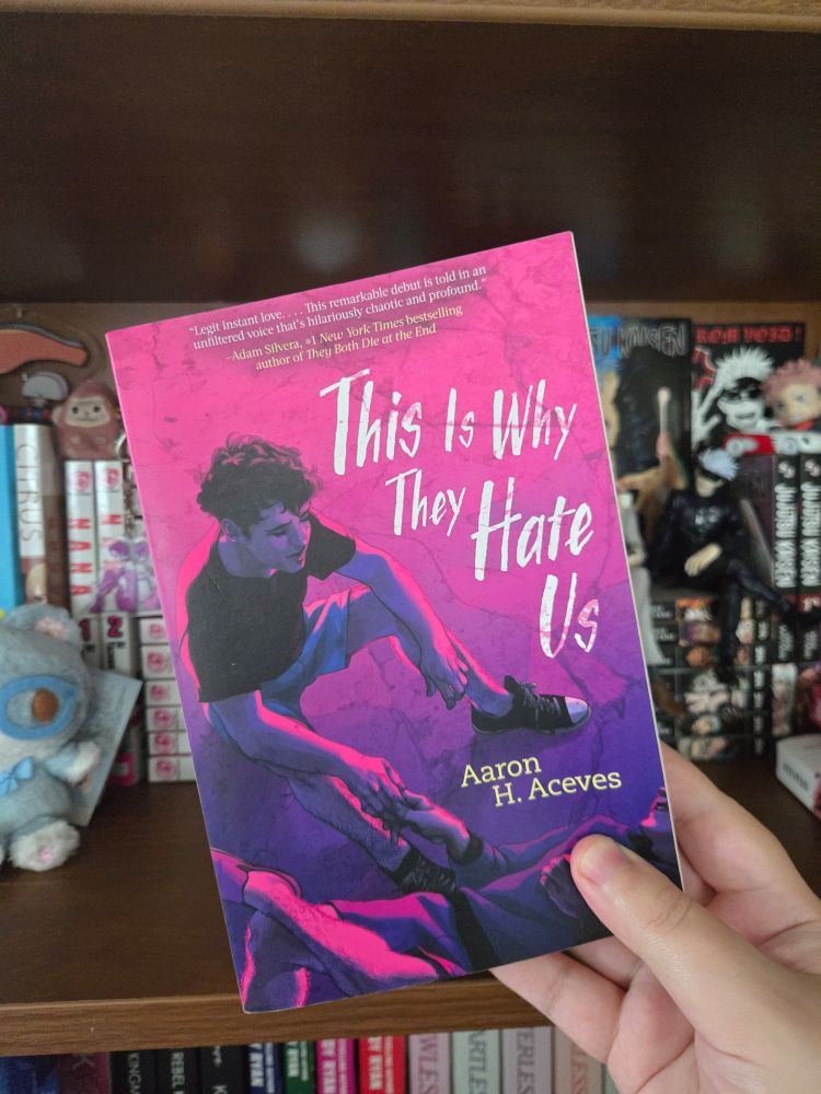 my right hand holding This Is Why They Hate Us by Aaron H. Aceves in front of my bookshelf