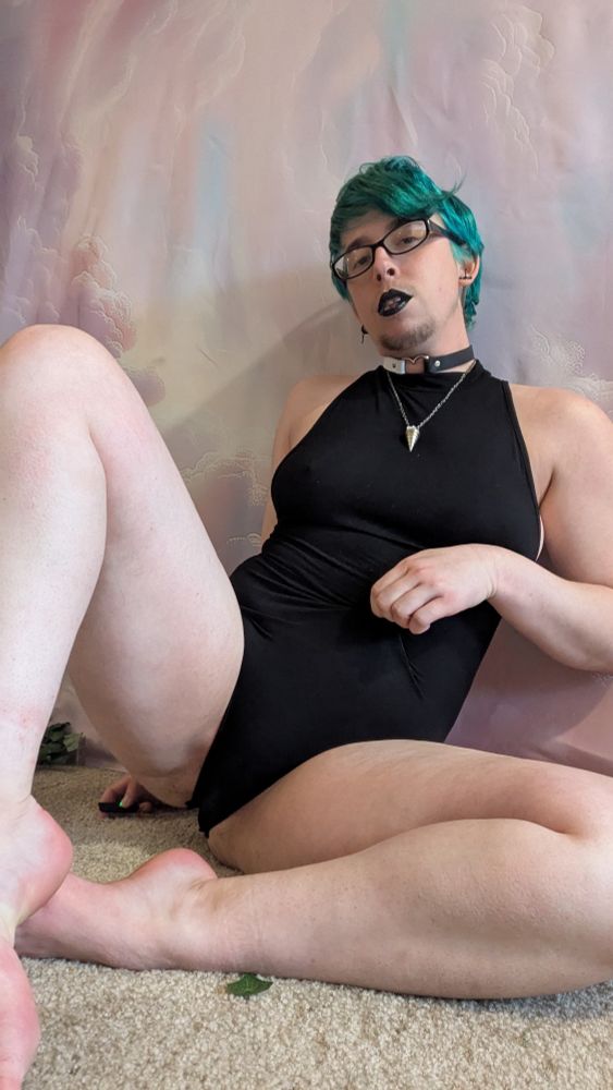 A person with blue hair and a black bodysuit sits on the floor with their bare legs out