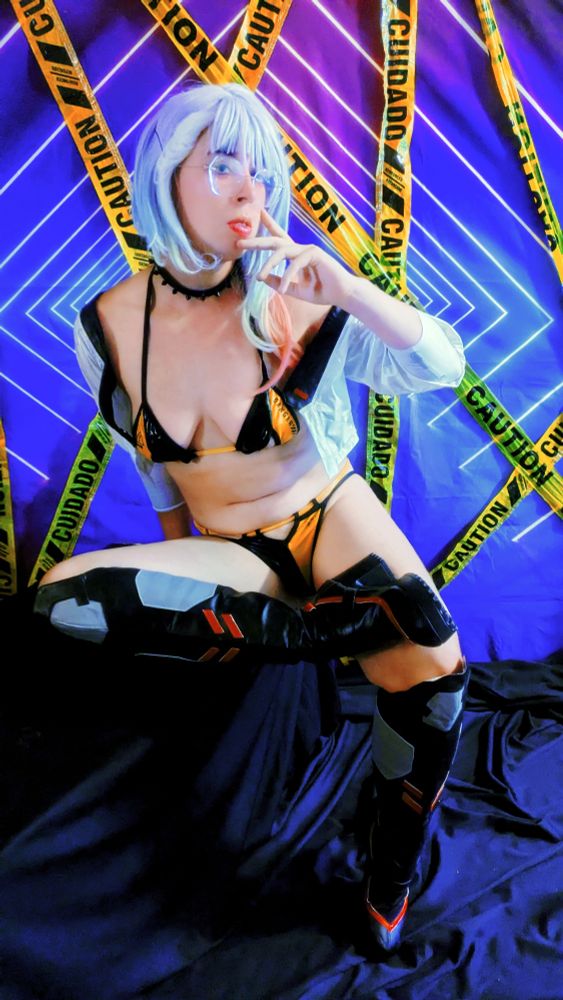 A cosplayer dressed in a yellow and black bikini with a cropped white coat.  They are also wearing cyberpunk style boots while sitting on a black box in front of a caution taped symmetrical backdrop