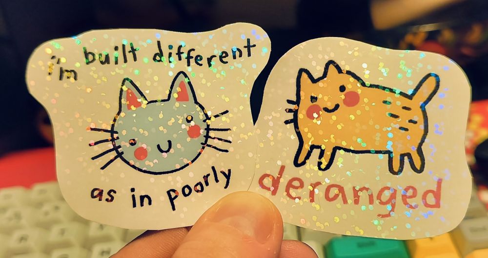 Photo of two sparkly stickers with white backgrounds. The first one has a wonky doodle of a light blue cats head. There is a text "I'm built different, as in poorly" around it. The second one is a yellow thick cat doodle with reddish text under it that says "deranged" both cats are smiling and blushing